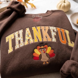 May include: A brown long-sleeve sweatshirt featuring the word THANKFUL in large, multi-patterned yellow and orange letters. Below the text is an embroidered turkey wearing a hat. The text DThuy DECOR SINCE 2024 is printed in small white letters.