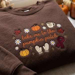 Pode incluir: Uma camisola de gola redonda castanho-escura com bordados de temática outonal, incluindo abóboras cor de laranja, margaridas brancas, uma caneca de café com um coração, um cachecol bordô e luvas beges, com o texto "take me to the pumpkin patch" em laranja.