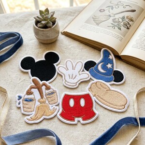 Op de afbeelding: Een verzameling geborduurde stoffen patches met een zwart Mickey Mouse-silhouet, een witte handschoen, een blauwe tovenaarshoed met sterren en maan, een beige bezem met twee emmers, een rode broek met witte knopen en een beige schoenvorm.