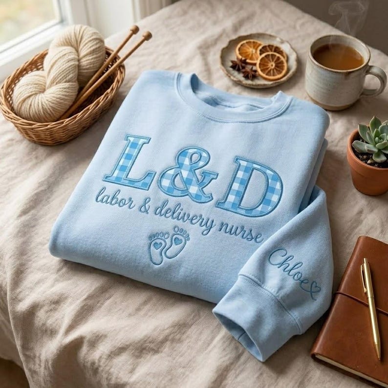 May include: A light blue sweatshirt with the embroidered text "L&D labor & delivery nurse" and baby footprints. The letters "L&D" are in a blue gingham pattern. The sleeve has the name "Chloe" embroidered on it.