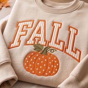 Op de afbeelding: Een beige sweater met ronde hals en het woord FALL geborduurd in grote blokletters met een wit en beige verticaal streeppatroon en oranje randen. Onder de tekst staat een geborduurde oranje pompoen met witte stippen.