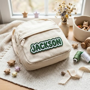 May include: A cream-colored canvas backpack featuring a green and white embroidered patch with the name JACKSON centered on the front pocket. The backpack has a zippered closure and a structured design, presented in a neutral, light-toned fabric.