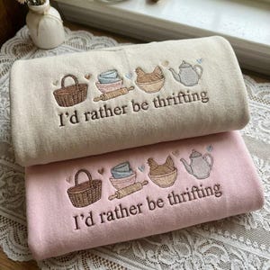 May include: Two embroidered rectangular towels, one beige and one pink, stacked on top of each other. Each towel features a basket, bowls, rolling pin, chicken, and teapot design with the text "I'd rather be thrifting".