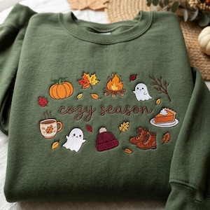 Pode incluir: Uma camisola de gola redonda verde-azeitona com bordados de tem&aacute;tica outonal, incluindo uma ab&oacute;bora, uma fogueira, fantasmas, um gorro, botas, uma fatia de tarte e uma caneca, com o texto "cozy season" em cursivo castanho no centro.
