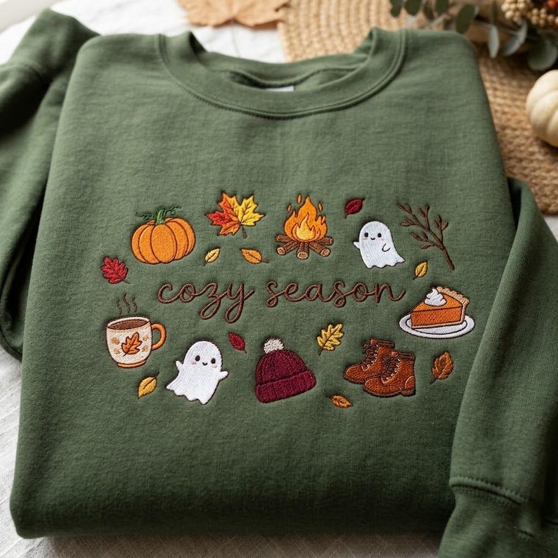 Pode incluir: Uma camisola de gola redonda verde-azeitona com bordados de tem&aacute;tica outonal, incluindo uma ab&oacute;bora, uma fogueira, fantasmas, um gorro, botas, uma fatia de tarte e uma caneca, com o texto "cozy season" em cursivo castanho no centro.