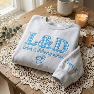 May include: A light gray sweatshirt with blue embroidered text and accents. The text reads "L&D labor & delivery nurse" with blue gingham-patterned letters. The sleeve cuff is embroidered with the name "Emma" and a heart.