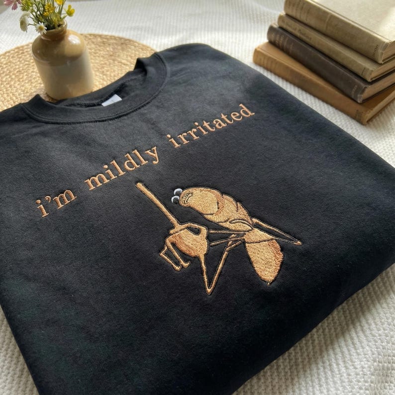 Może przedstawiać: Czarna bluza z haftowanym napisem "I'm mildly irritated" nad szczeg&oacute;łową ilustracją osy trzymającej małe widły. Tekst i osa są w kolorze jasnobrązowym.
