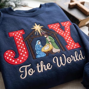 May include: A navy blue sweatshirt featuring embroidered text that reads JOY with the letter O replaced by a nativity scene inside a wooden stable under a gold star. Below the graphic, the text To the World is embroidered in tan script.