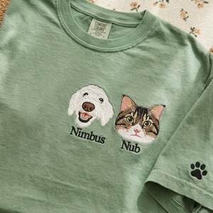 May include: A sage green t-shirt featuring embroidered portraits of a white dog and a brown tabby cat. The dog is labeled Nimbus and the cat is labeled Nub in black thread. A small black paw print is embroidered on the sleeve of the shirt.