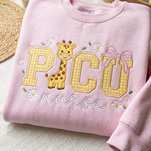 Op de afbeelding: Roze sweatshirt met geborduurde tekst en afbeeldingen. Het woord "PICU" staat in gele ruitjesletters, met een giraf in het midden. Het woord "nurse" is eronder in cursief geborduurd, met madeliefjesaccenten.