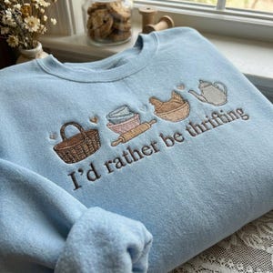 May include: A light blue sweatshirt with embroidered baking-themed images, including a basket, bowls, rolling pin, and teapot. The phrase "I'd rather be thrifting" is embroidered in brown. The sweatshirt is made of a soft material.