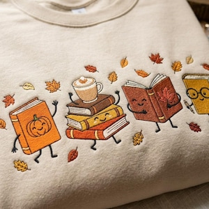 May include: A beige crewneck sweatshirt featuring an embroidered design of anthropomorphic books and a coffee mug. The scene includes a pumpkin-adorned book, a stack of three books with a mug on top, an open book, and a book wearing glasses.