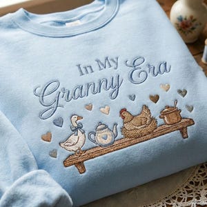 May include: Light blue sweatshirt with embroidered text "In My Granny Era". The design includes a duck with a bow, a teapot, a hen, and a pot on a wooden shelf, with heart accents. A cozy, vintage-inspired design.