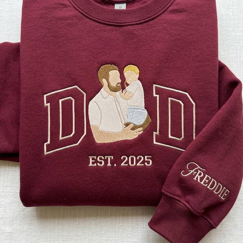 May include: Burgundy sweatshirt with embroidered design. Features the word "DAD" in a collegiate font, an illustration of a father holding a child, and the text "EST. 2025". The sleeve is embroidered with the name "Freddie".