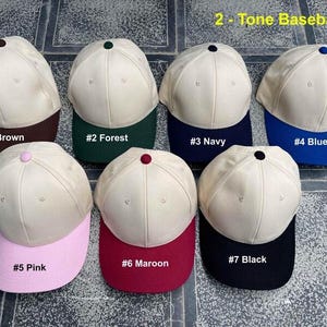 May include: A collection of seven two-tone baseball caps. Each cap has a cream-colored crown and a colored brim. The brims are brown, forest green, navy, blue, pink, maroon, and black. The text "2 - Tone Baseball Cap" is at the top.