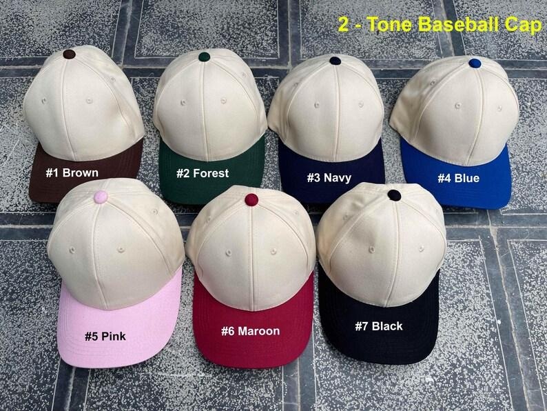 May include: A collection of seven two-tone baseball caps. Each cap has a cream-colored crown and a colored brim. The brims are brown, forest green, navy, blue, pink, maroon, and black. The text "2 - Tone Baseball Cap" is at the top.