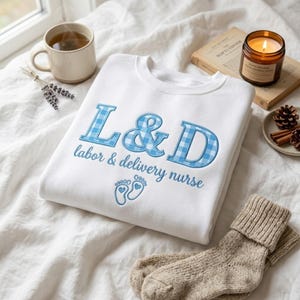 May include: A white sweatshirt with the embroidered text "L&D labor & delivery nurse" in blue gingham lettering. The design includes baby footprints. A cup of tea, a lit candle, and a pair of socks are also in the image.
