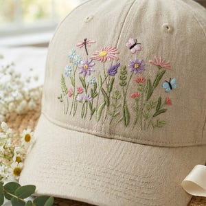 Op de afbeelding: Beige baseballpet met een geborduurd bloemenontwerp. De borduursels tonen kleurrijke bloemen, groene bladeren, een libel en vlinders. De pet is gemaakt van een textuurstof.