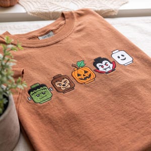 May include: A rust-colored t-shirt featuring an embroidered row of five Halloween-themed toy figures. The designs include a green monster, a brown werewolf, an orange jack-o'-lantern, a vampire with a red cape, and a plain white toy head.