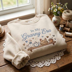 May include: Beige sweatshirt with the embroidered phrase "In My Granny Era" in blue script. The design includes a goose, teapot, chicken, and pot on a wooden shelf, with heart accents.