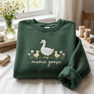 May include: A dark green sweatshirt with an embroidered design of a white goose, two yellow ducklings, and the words "mama goose" in cursive. The design includes small white flowers. The sweatshirt has a crew neck and long sleeves.