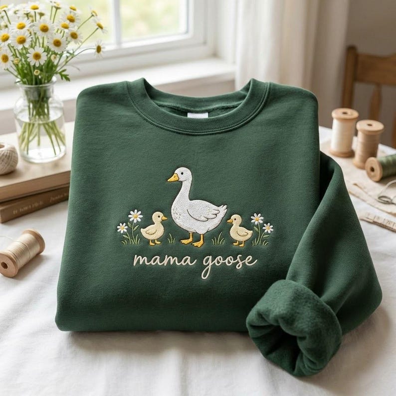 May include: A dark green sweatshirt with an embroidered design of a white goose, two yellow ducklings, and the words "mama goose" in cursive. The design includes small white flowers. The sweatshirt has a crew neck and long sleeves.