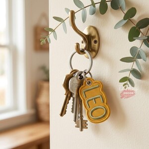 Op de afbeelding: Een messingkleurige metalen wandhaak houdt een zilveren metalen sleutelhanger met diverse sleutels en een mosterdgeel stoffen label met de geborduurde tekst LEO in een saliegroene omlijning, geaccentueerd door een takje met groene eucalyptusbladeren.