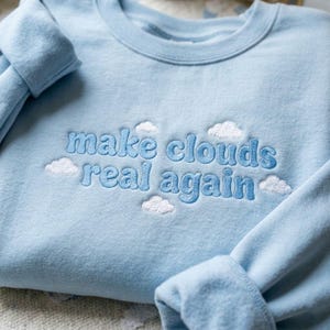 May include: A light blue sweatshirt with the embroidered phrase "make clouds real again" in a lighter blue font. White cloud designs are embroidered around the text. The sleeves are folded.