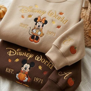 May include: A beige sweatshirt featuring embroidered text Disney World, EST. 1971, and Minnie Mouse holding a mug, surrounded by autumn leaves. A dark brown sweatshirt with embroidered text Disney World, EST. 1971, and Mickey Mouse holding a pumpkin.