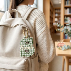 May include: A beige fabric backpack featuring a hanging keychain accessory. The keychain is a green and white gingham patterned letter S with the name Sarah embroidered in white script across the center, attached by a gold metal carabiner.