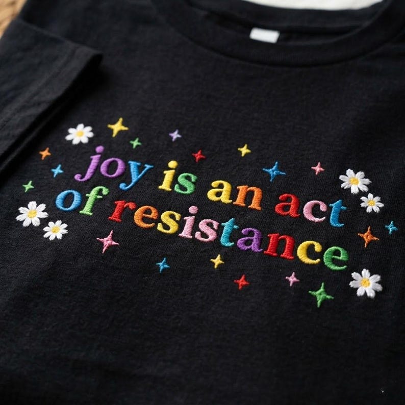 Pu&ograve; includere: T-shirt nero con testo ricamato "joy is an act of resistance" in colori arcobaleno. Il design include stelle e margherite ricamate, creando un'estetica colorata e allegra.