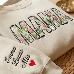 Op de afbeelding: Cr&egrave;mekleurige sweatshirt met het geborduurde woord "MAMA" in zwarte omtrek, gevuld met roze tulpen, witte madeliefjes en groene bladeren. De mouw heeft geborduurde namen en een rood hart.