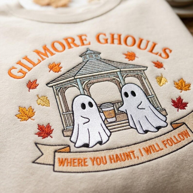 May include: A beige sweatshirt featuring an embroidered design of a gazebo, two white ghosts, and falling autumn leaves. The text reads GILMORE GHOULS at the top and WHERE YOU HAUNT, I WILL FOLLOW on a banner at the bottom in orange thread.
