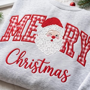May include: A light grey sweatshirt featuring the embroidered red text MERRY with a red and white gingham pattern. A Santa Claus face with a red hat and white beard replaces the letter E. The word Christmas is embroidered below in red script.