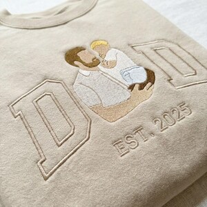 May include: Beige sweatshirt with embroidered design. Features the word "DAD" in a collegiate font, with an illustration of a father and child. The text "EST. 2025" is embroidered below the word "DAD".