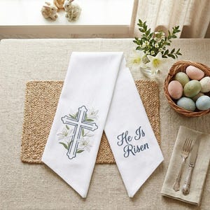 May include: White linen Easter towels with embroidered cross and lily design. One towel has the text "He Is Risen" in blue script. The towels are displayed with a basket of pastel Easter eggs, flowers, and decorative bunnies.