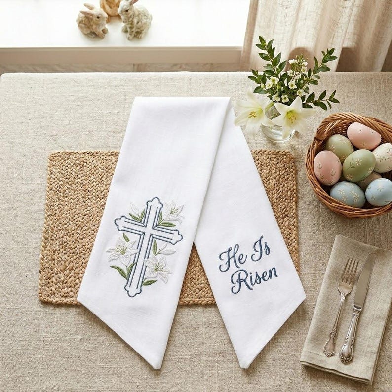 May include: White linen Easter towels with embroidered cross and lily design. One towel has the text "He Is Risen" in blue script. The towels are displayed with a basket of pastel Easter eggs, flowers, and decorative bunnies.