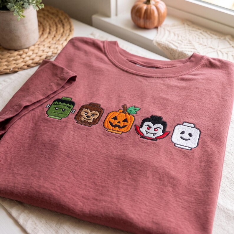 May include: A dusty rose crewneck t-shirt featuring five embroidered block-style character heads in a horizontal row. The designs include a green monster, a brown werewolf, an orange jack-o'-lantern, a vampire, and a white smiling ghost figure.