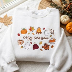 Pode incluir: Uma camisola branca de gola redonda com bordados coloridos de tem&aacute;tica outonal, incluindo uma ab&oacute;bora, fogueira, fantasmas, uma caneca, um gorro, botas e uma fatia de tarte, com o texto "cozy season" em letras cursivas castanhas.