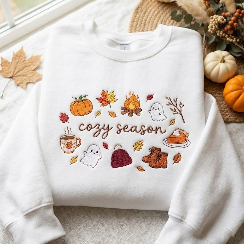 Pode incluir: Uma camisola branca de gola redonda com bordados coloridos de tem&aacute;tica outonal, incluindo uma ab&oacute;bora, fogueira, fantasmas, uma caneca, um gorro, botas e uma fatia de tarte, com o texto "cozy season" em letras cursivas castanhas.