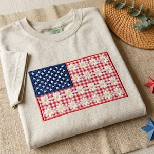 May include: A beige crew neck t-shirt with an embroidered American flag design. The flag's blue field has white stars, while the stripes are replaced with a red and white gingham pattern and white daisy flowers.