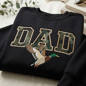 May include: Black sweatshirt with the word "DAD" in a camouflage design. Below the text is an embroidered image of a flying duck with brown, green, and blue feathers. The sweatshirt is a crew neck style.