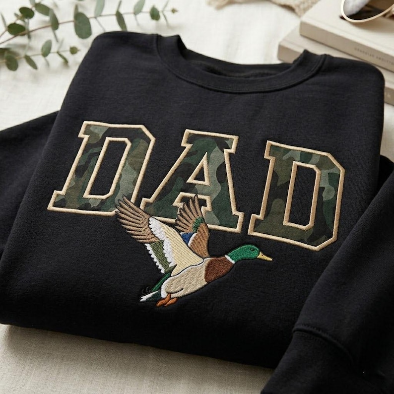 May include: Black sweatshirt with the word "DAD" in a camouflage design. Below the text is an embroidered image of a flying duck with brown, green, and blue feathers. The sweatshirt is a crew neck style.