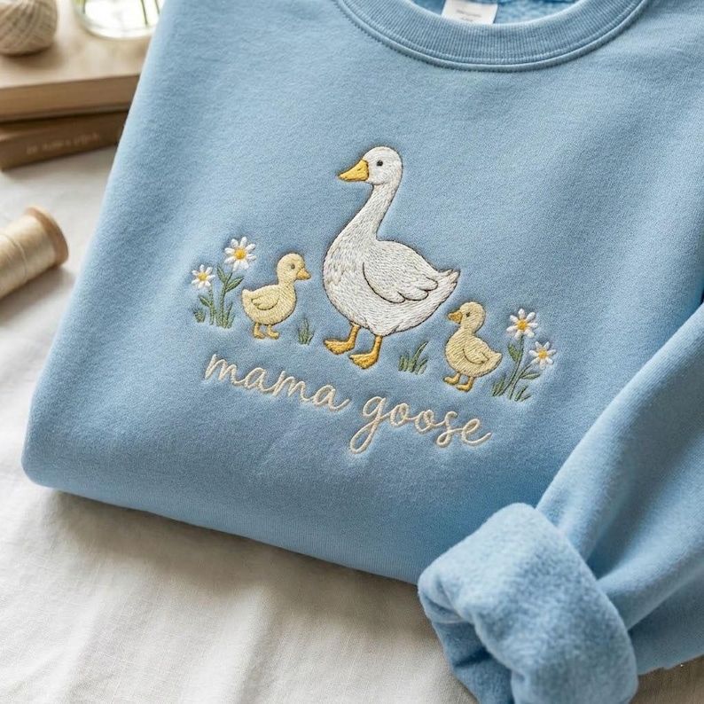 May include: Light blue sweatshirt with an embroidered design featuring a white goose, two yellow goslings, and daisies. The words "mama goose" are embroidered below the design. The sweatshirt has a crew neck and cuffed sleeves.