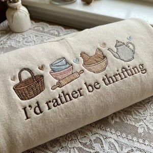 May include: Cream-colored sweatshirt embroidered with kitchen-themed illustrations. Features a basket, bowls, rolling pin, chicken, and teapot. The phrase "I'd rather be thrifting" is embroidered in brown script. The design includes small heart accents.