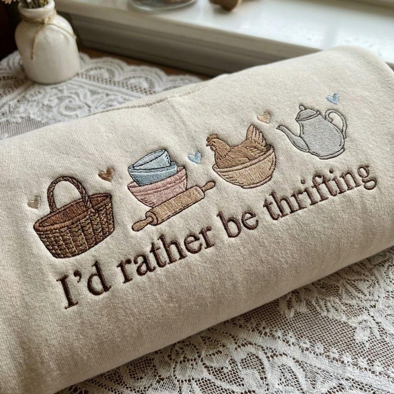 May include: Cream-colored sweatshirt embroidered with kitchen-themed illustrations. Features a basket, bowls, rolling pin, chicken, and teapot. The phrase "I'd rather be thrifting" is embroidered in brown script. The design includes small heart accents.