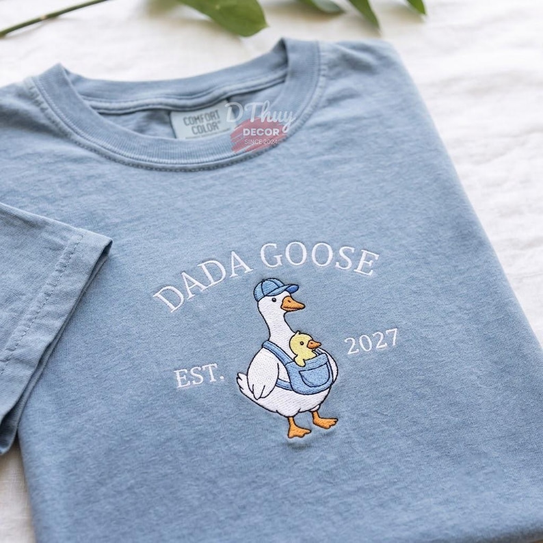 May include: A dusty blue t-shirt featuring an embroidered graphic of a white goose wearing a blue baseball cap and a blue baby carrier. A small yellow gosling sits inside the carrier. The text DADA GOOSE and EST. 2027 are embroidered in white.