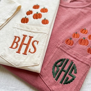 Może przedstawiać: Kremowy t-shirt z pomarańczowymi haftowanymi dyniami i pomarańczowym monogramem BHS na kieszeni, obok brudnor&oacute;żowego t-shirtu z pomarańczowymi haftowanymi dyniami i ciemnozielonym okrągłym monogramem BHS na kieszeni.