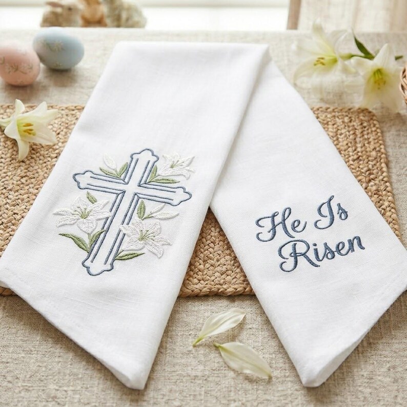 May include: Two white linen towels with embroidered designs. One towel features a blue cross with white lilies and green leaves. The other towel has the phrase "He Is Risen" embroidered in blue script. Easter themed.