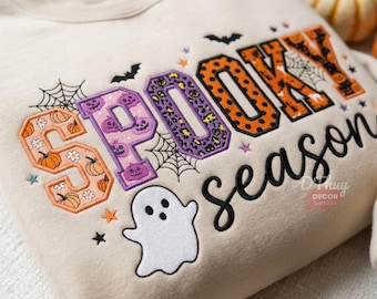 Op de afbeelding: Een beige sweater met ronde hals en de geborduurde tekst SPOOKY season. De letters zijn gevuld met oranje pompoenpatronen, paarse spookontwerpen en oranje luipaardprint. Een wit spookje, zwarte spinnenwebben en vleermuizen zijn ook geborduurd.